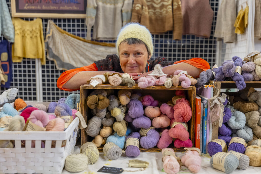 la teinturière sur son stand pendant un salon, devant et derrière des fils et des créations tricotées colorées